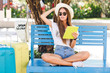 © Look! - Cute girl sitting on a blue bench and playing on a tablet in yellow case. She wears denim shorts, white t-shirt, dark sunglasses, white sneakers and straw hat. She sits with two suitcases next to her.