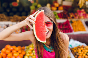 Closeup portrait of funny girl in pink sunglasses holding slice of watermelon on half face on tropical fruits background.