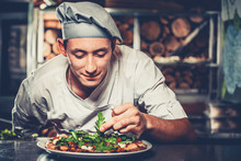 Food Concept. Preparing Traditional Italian Pizza. Young Smiling Chef In White Uniform And Gray Hat Decorate Ready Dish With Green Rucola Herbs In Interior Of Modern Restaurant Kitchen. Ready To Eat.