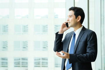 Wall Mural - Business man talking smart phone with happy smiling face standing by windows with city view, inside office building background, businessman on smart phone