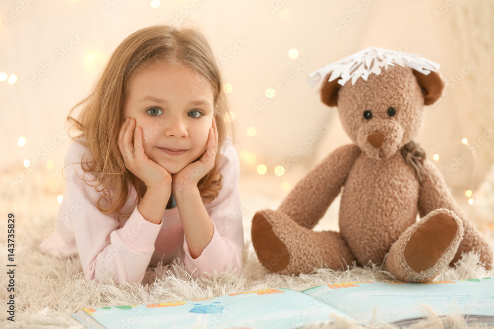 Cute little girl with teddy bear reading book at home