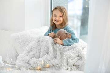 Cute little girl with teddy bear sitting on window sill at home