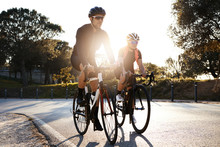 Handsome Bearded Professional Male Cyclist Riding His Racing Bicycle In The Morning Together With His Girlfriend, Both Wearing Protective Helmets And Eyeglasses, Sun Shining Through Between Them