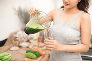 Wall Mural - Smiling asian woman making smoothie with fresh vegetables in the blender in kitchen at home.