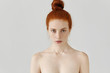 Isolated portrait of beautiful young Caucasian redhead woman with hair bun and perfect clean skin with freckles standing at grey studio wall with shoulders naked, having serious facial expression