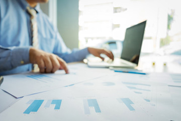 Wall Mural - Handsome young businessman working with laptop while sitting on the desk in office and financial statistic graph, blurred background