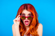 Surprised shocked girl with beautiful red curly long hair holding sunglasses and open mouth while standing on blue background