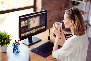 Female graphic designer sitting near screen and graphic tablet at the desktop in workstation and have a break with cup of coffee, back view