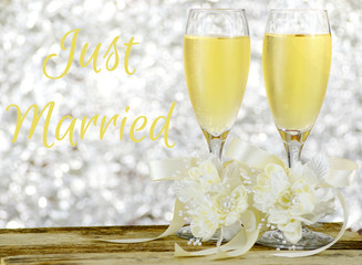 A pair of glasses filled with sparkling bubbly champagne and decorated with ivory colored satin ribbons and silk flowers on a wooden table in front of a sparkly, glittering silver bokeh background