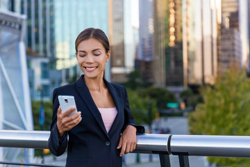 Wall Mural - Woman sms texting using app on smart phone in city business district. Young business woman using smartphone smiling happy wearing suit jacket outdoors. Urban female professional in her 20s.