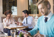 © phocal Media - Man using digital tablet in restaurant with people in background.
