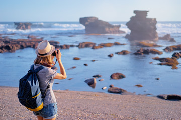 Wall Mural - Traveling and photography. Young woman with camera and backpack taking picture on the sea beach.