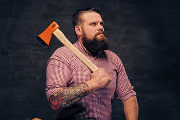 Wall Mural - Bearded lumberjack male with tattoos on arms, sits on a wooden box and holds the axe.