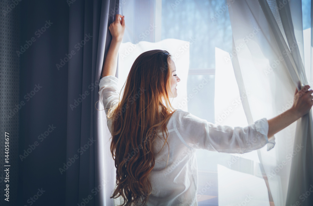 Pretty woman in modern apartment opening window curtains after wake up - obrazy, fototapety, plakaty 