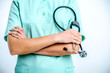 © daria_serdtseva - A female doctor holds a black phonendoscope in her hands. The doctor is wearing a green surgical suit.