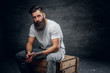 © Fxquadro - Bearded male with tattoo on arm dressed in a white t shirt sits on a wooden box.