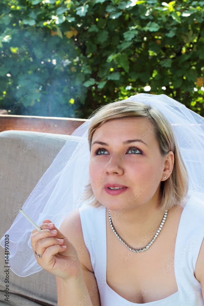 Young nervous sad bride in a white wedding dress smoking a cigarette  outdoors against a background of green foliage Stock Photo