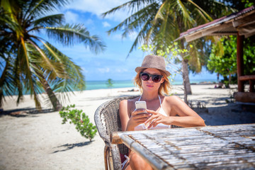 Young beautiful woman with mobile phone on the shore of the tropical sea in a cafe. Communication and internet in travel concept