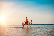 Woman And Horse On The Background Of Sky And Water. Girl Model On Horseback On The Beach By The Sea At Sunset, Backlit In Sunshine. A Positive Summer Time Scene.