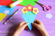 Little child made paper crafts for mother's day or birthday. Child holds a paper bouquet in hands. Easy and beautiful gift for mom. Scissors, glue stick, flowers templates, pencil on a wooden table