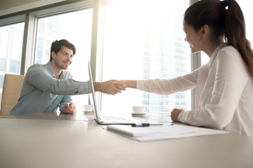 Wall Mural - Businesswoman and businessman shaking hands over the table with laptop and papers at the office, pleasant business meeting, starting negotiation, conducting job interview