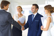 © volha_r - Two confident businessmen shaking hands and smiling while standing at office together with group of  colleagues.