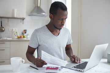 Concentrated young african american student using online banking application with hand on touchpad, looking at screen of his laptop, trying to make payment for university education. Financial problems