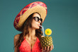 young beautiful woman in bikini, sunglasses and sombrero drinking tropic cocktail isolated on blue background, close-up