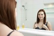 © Saksoni - morning and people concept - young woman looking to mirror at home bathroom