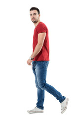 Wall Mural - Side view of young relaxed casual man walking and looking at camera. Full body length portrait isolated over white studio background.