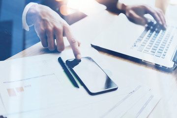 Man working on modern mobile phone and laptop at sunny office and pointing finger to home button of smartphone.Horizontal, blurred background, visual effects.