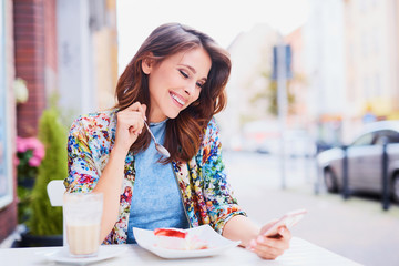 Happy woman with smartphone eating cake at outdoors cafe