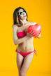 © bluebeat76 - Beautiful brunette woman in pink bikini playing with red heart shape balloon isolated on yellow background. Toned Image.