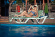 © satyrenko - Beautiful girl is sunbathing in swimsuit with pleasure. She is lying near a swimming pool. The lady is enjoying the sun and smiling. She is wearing sunglasses and a hat