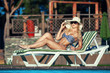© satyrenko - Beautiful girl is sunbathing in swimsuit with pleasure. She is lying near a swimming pool. The lady is enjoying the sun and smiling. She is wearing sunglasses and a hat