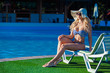 © satyrenko - Beautiful young girl is sitting on the chaise-longue with pleasure near a swimming pool. She is wearing beautiful swimsuit, sunglasses and a hat