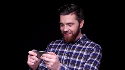 Wall Mural - Excited young bearded man playing games on smartphone isolated over black background