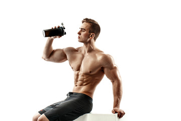 Muscular man with protein drink in shaker over white background sitting on cube. Studio shot.