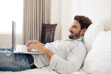 Happy handsome man using laptop. Lying on bed in bedroom.