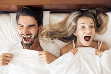 Top View Of Beautiful Young Woman And Her Man Showing Surprise And Looking At Camera While Lying In Bed Under Blanket