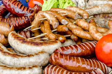 assorted sausages grilled on skewers on a large plate, tomatoes on the grill. Street festival food.