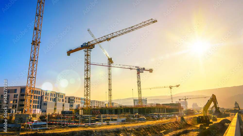 Large construction site including several cranes working on a building complex, with clear blue sky and the sun - obrazy, fototapety, plakaty 