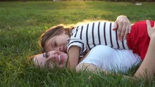 Mother And Daughter Lying On The Lawn. Daughter Kiss Her Mother On The Cheek. Family In The City Park Outdoors. Happiness Of Motherhood And Childhood. On The Sunset