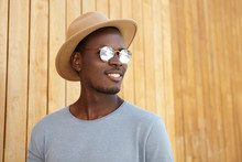 Cheerful Carefree Young Dark-skinned Man Wearing Mirrored Lens Shades And Trendy Hat Smiling Happily, Rejoicing At Warm Sunny Weather, Posing Isolated At Wooden Wall With Copy Space For Your Content