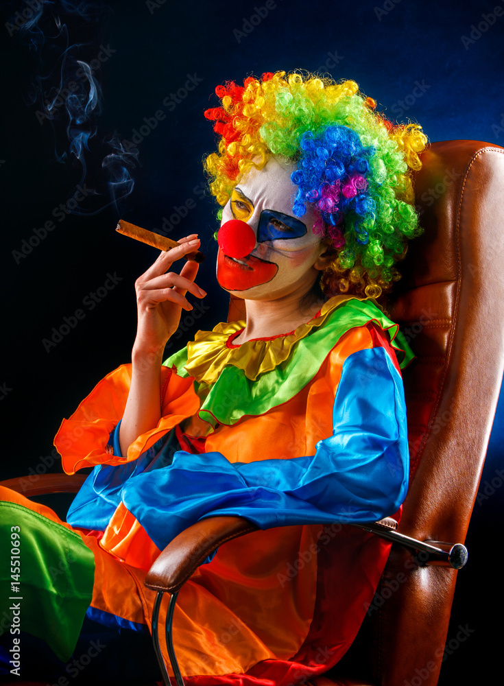 crazy cigarette Mad clown on black background. Woman is smoking cigar. Portrait of crazy  female with angry look after performance sits in leather armchair. Stock  Photo