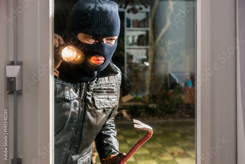 Fotomural Masked burglar with flashlight and crowbar looking into glass window of house