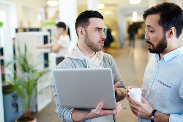 Canvas Print - Two modern business people discussing work project standing with laptop in open office space