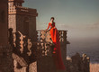 © kharchenkoirina - Beautiful girl sitting on the balcony of the castle. The princess has a red dress with a long train. The backdrop of the sky and the sea.