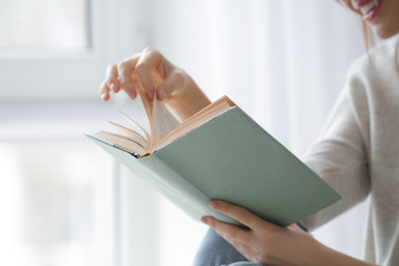 Beautiful young woman reading book near window at home, closeup