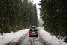 Red Car With Roof Rack Driving On A Forest Road In Winter, Heavy Snowfall. The Concept Of Traveling By Car